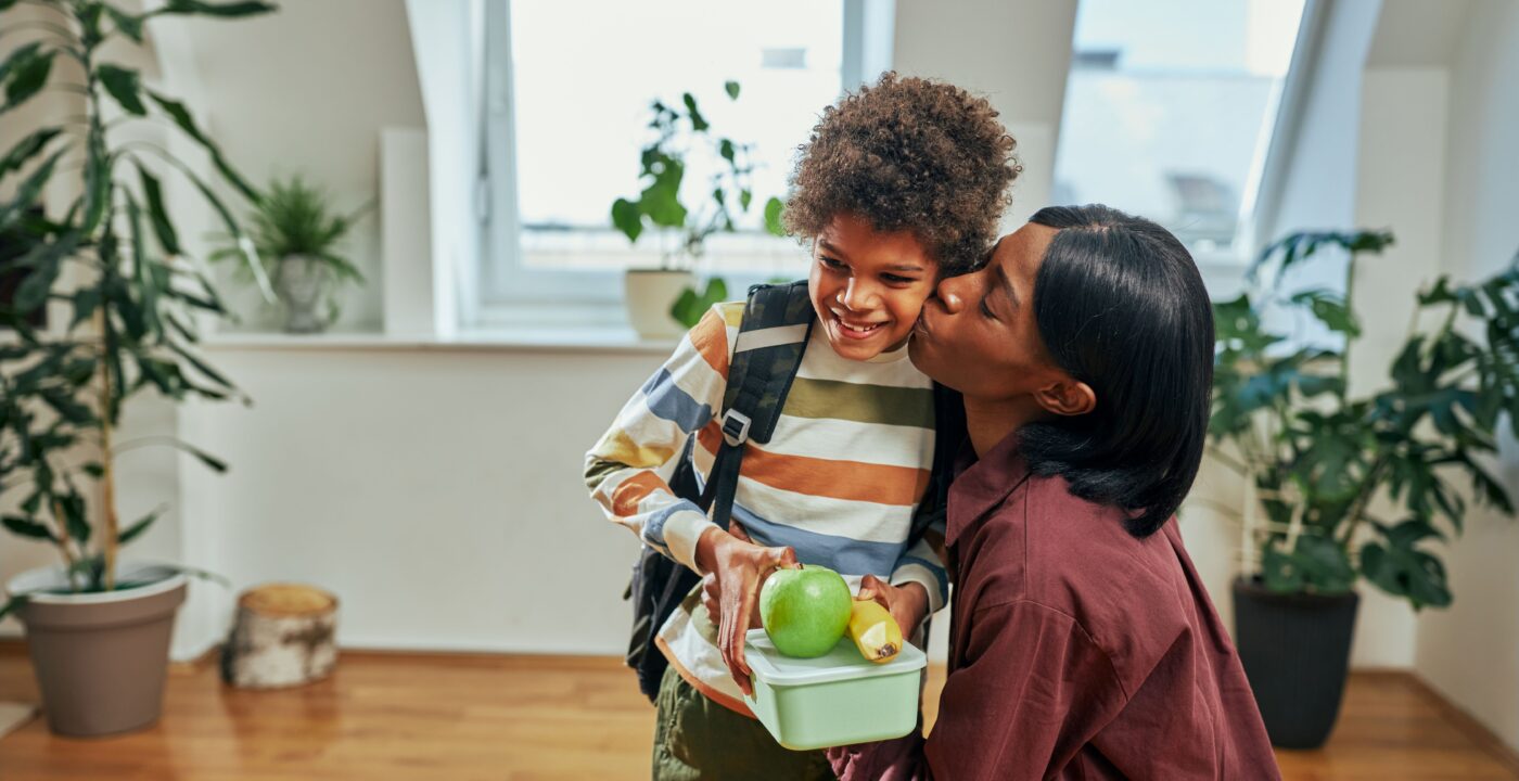 Child holding his lunch as his mom kisses him goodbye