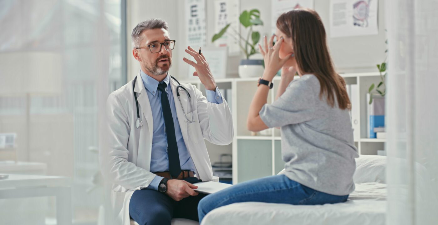 woman sits on exam table, pointing to her head while talking with doctor