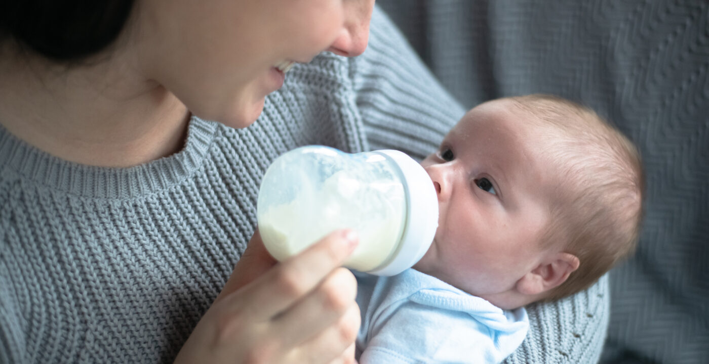 A Caucasian mother cradles her new infant baby boy in her arms as she bottle feeds him. She is leaning in close and smiling down at him as she holds the bottle propped up in his mouth. The baby is wearing a diaper shirt and the other is dressed casually in a sweater.