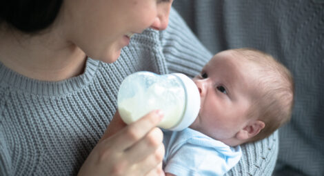 A Caucasian mother cradles her new infant baby boy in her arms as she bottle feeds him. She is leaning in close and smiling down at him as she holds the bottle propped up in his mouth. The baby is wearing a diaper shirt and the other is dressed casually in a sweater.