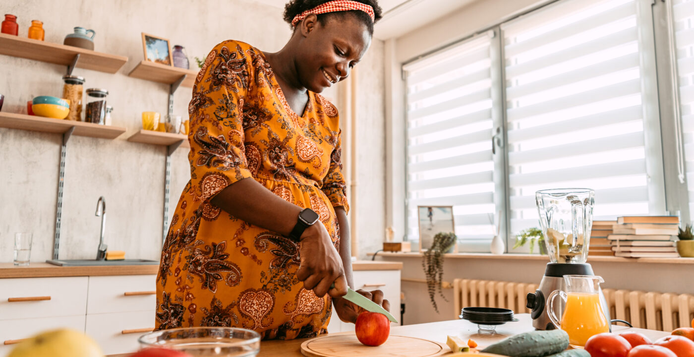 A woman slices a red apple on a cutting board in a bright home kitchen, with fresh fruits, vegetables, and a blender on the counter.
