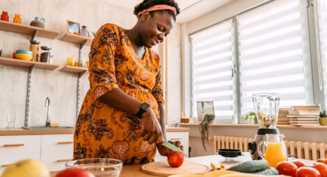 A woman slices a red apple on a cutting board in a bright home kitchen, with fresh fruits, vegetables, and a blender on the counter.