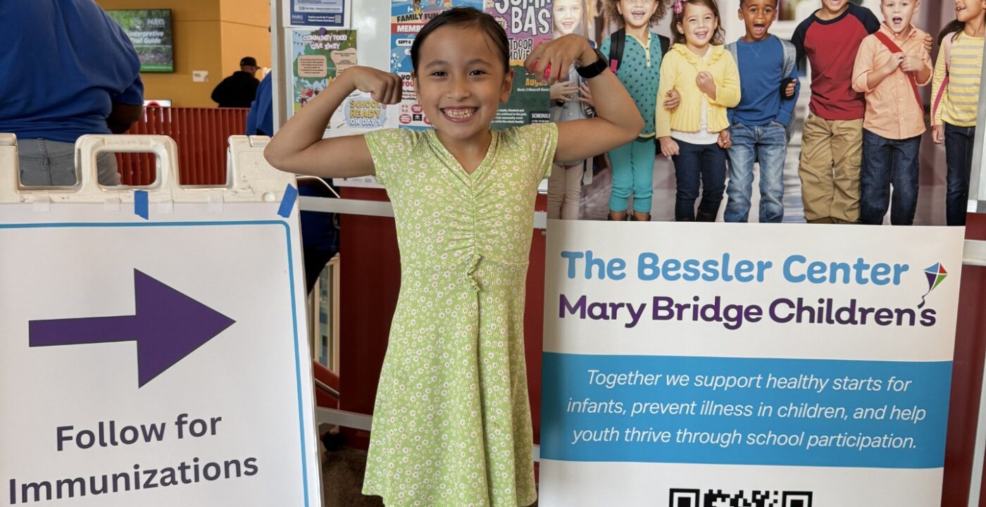 Child poses with arms flexed next to a sign for the Bessler Center at Mary Bridge Children’s and a directional sign for immunizations at an indoor event.
