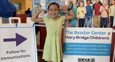 Child poses with arms flexed next to a sign for the Bessler Center at Mary Bridge Children’s and a directional sign for immunizations at an indoor event.