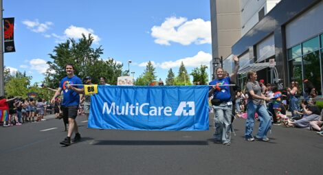 Parade participants carry a large blue banner reading “MultiCare” while walking down a street during a Pride event, with crowds watching on both sides.