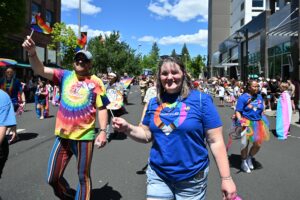 People walk in a Pride parade on a city street, wearing colorful clothing and waving small rainbow flags, with spectators lining the sidewalks.