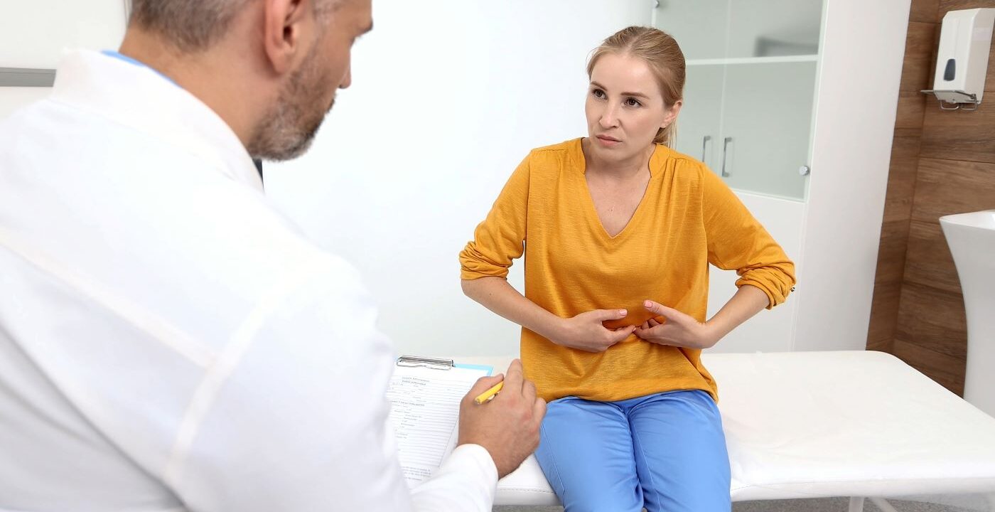 woman wearing orange shirt pressing on abdomen talking to doctor
