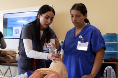 Instructor demonstrates proper use of a handheld airway device on a medical training mannequin while another participant in scrubs observes closely in a classroom setting, with a screen in the background showing intubation visuals.