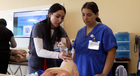 Instructor demonstrates proper use of a handheld airway device on a medical training mannequin while another participant in scrubs observes closely in a classroom setting, with a screen in the background showing intubation visuals.
