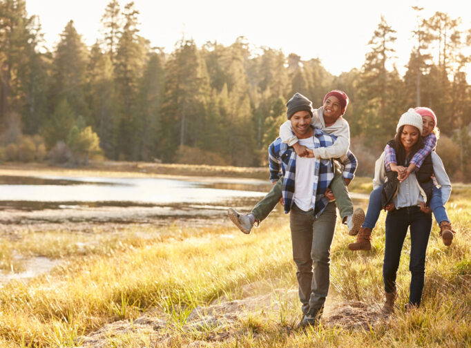 Family walks along side a creek on a fall day. The two kids riding on their mom and dad's backs.