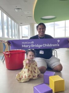 A small child sits on the floor of a hospital playroom while another person kneels behind them holding a purple “MultiCare Mary Bridge Children’s” ribbon; colorful foam blocks and a red bucket are nearby, with large windows in the background.