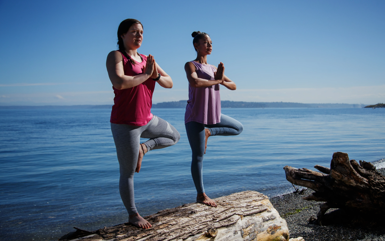Two women doing yoga on a log by the water