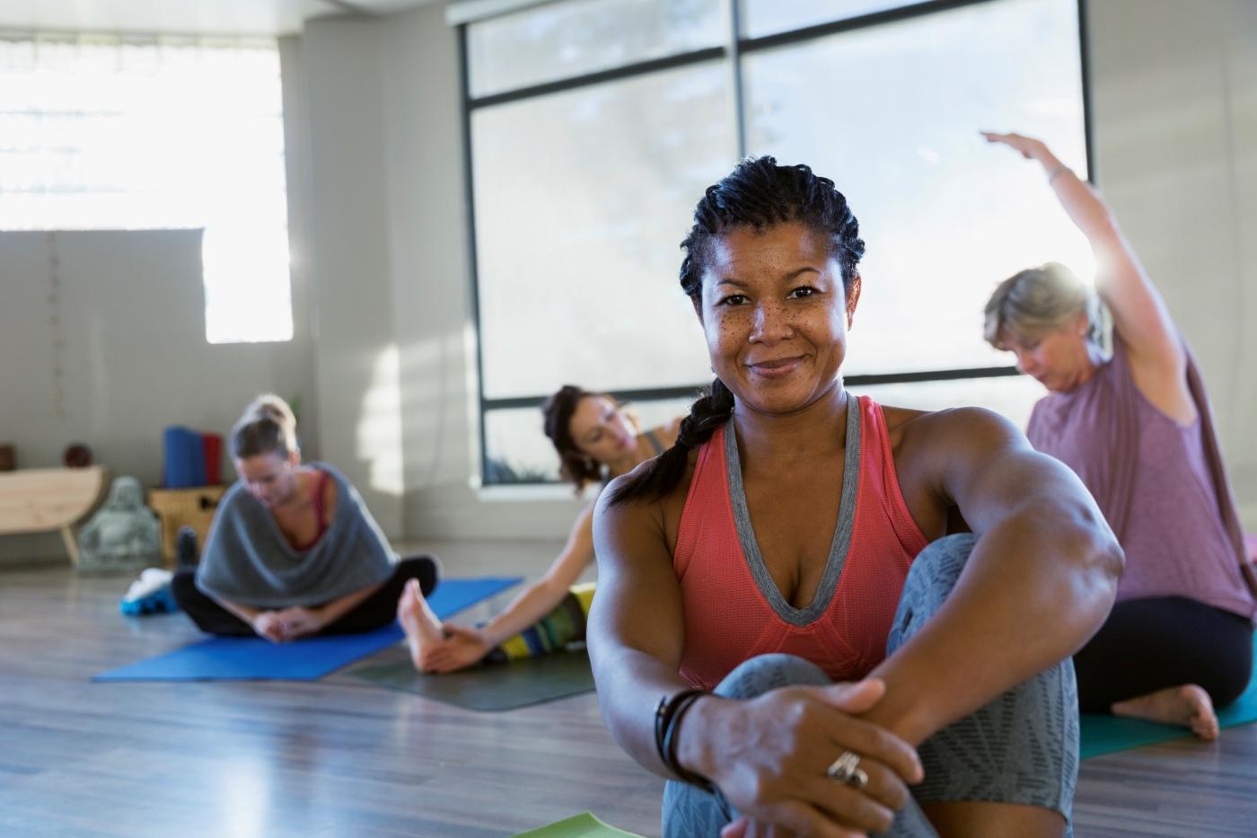 Woman smiles for camera while sitting on the floor in a bright room with lots of windows as other women stretch on mats behind her.