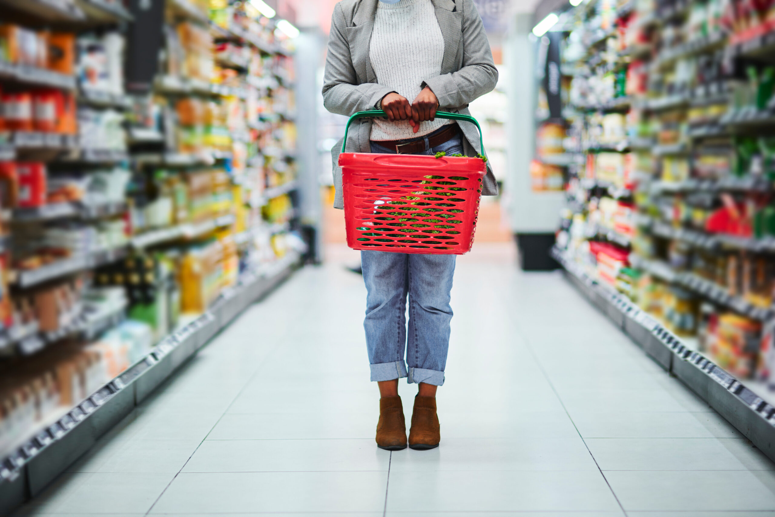 Woman pictured from waist down holding a shopping basket in a grocery store aisle