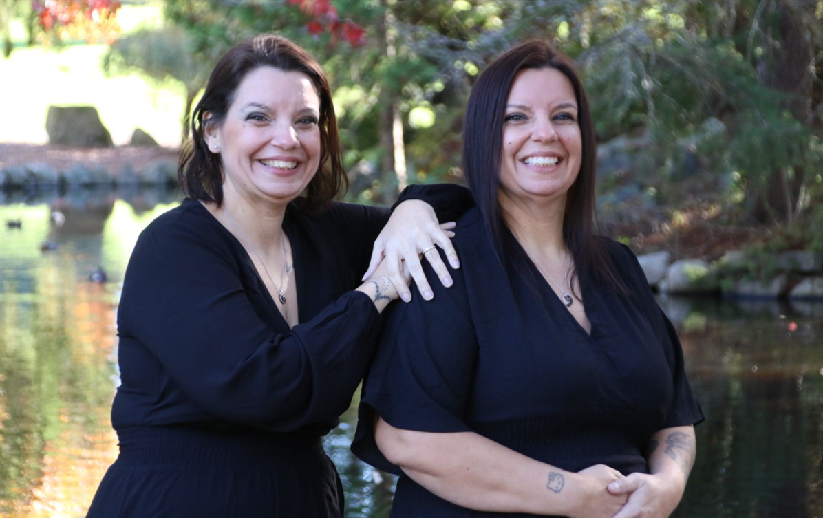 Twin women in black dresses and silver necklaces pose in front of a pond on a sunny day; one twin has short brown hair and the other has long dark brown hair