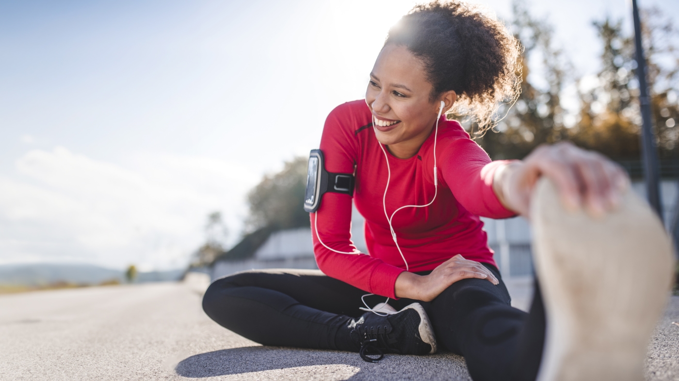 Woman in running gear sits on the pavement and stretches with a hand grabbing her shoe at the toes. It’s a sunny day and she has a big smile on her face.