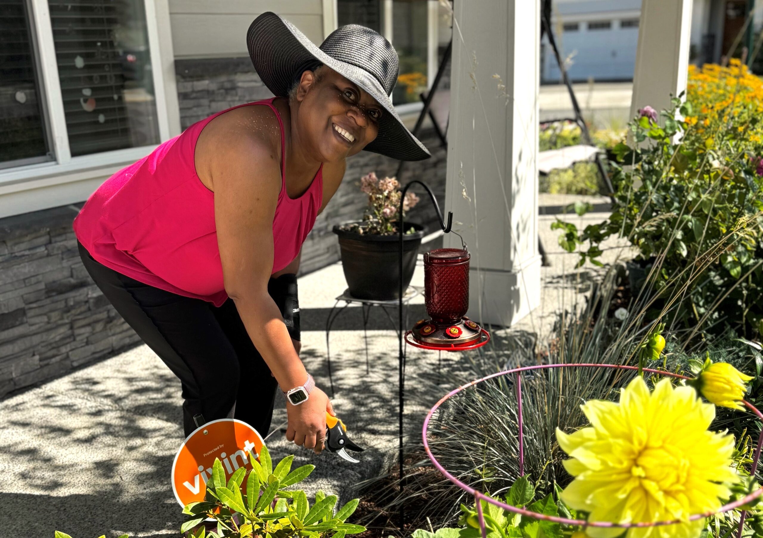 Woman in wide-brimmed hat holds plant clippers above a plant bed while smiling at the camera