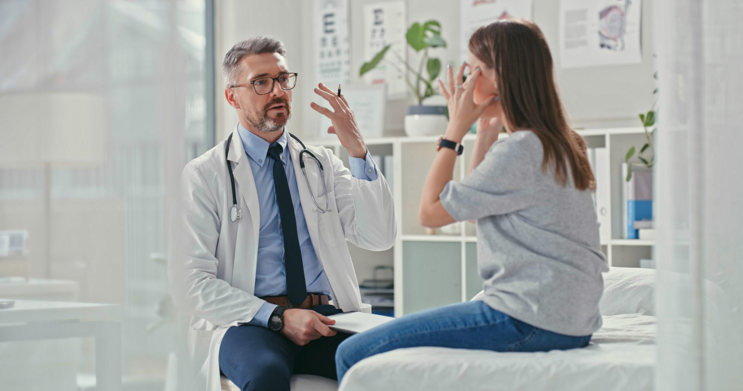 woman sits on exam table, pointing to her head while talking with doctor