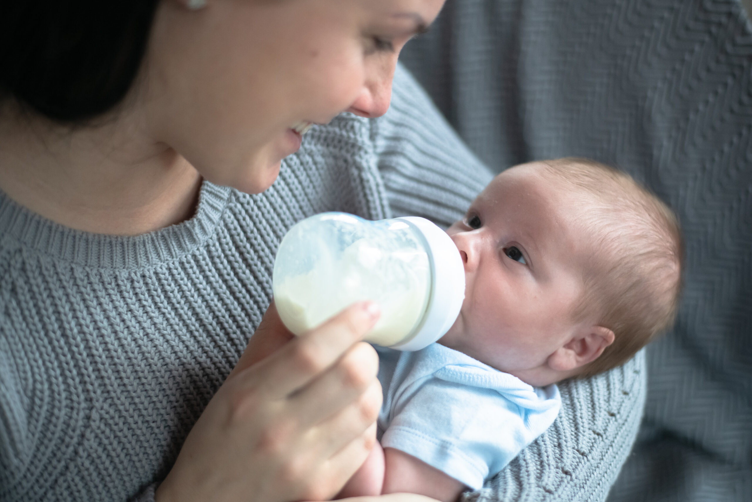 A Caucasian mother cradles her new infant baby boy in her arms as she bottle feeds him. She is leaning in close and smiling down at him as she holds the bottle propped up in his mouth. The baby is wearing a diaper shirt and the other is dressed casually in a sweater.