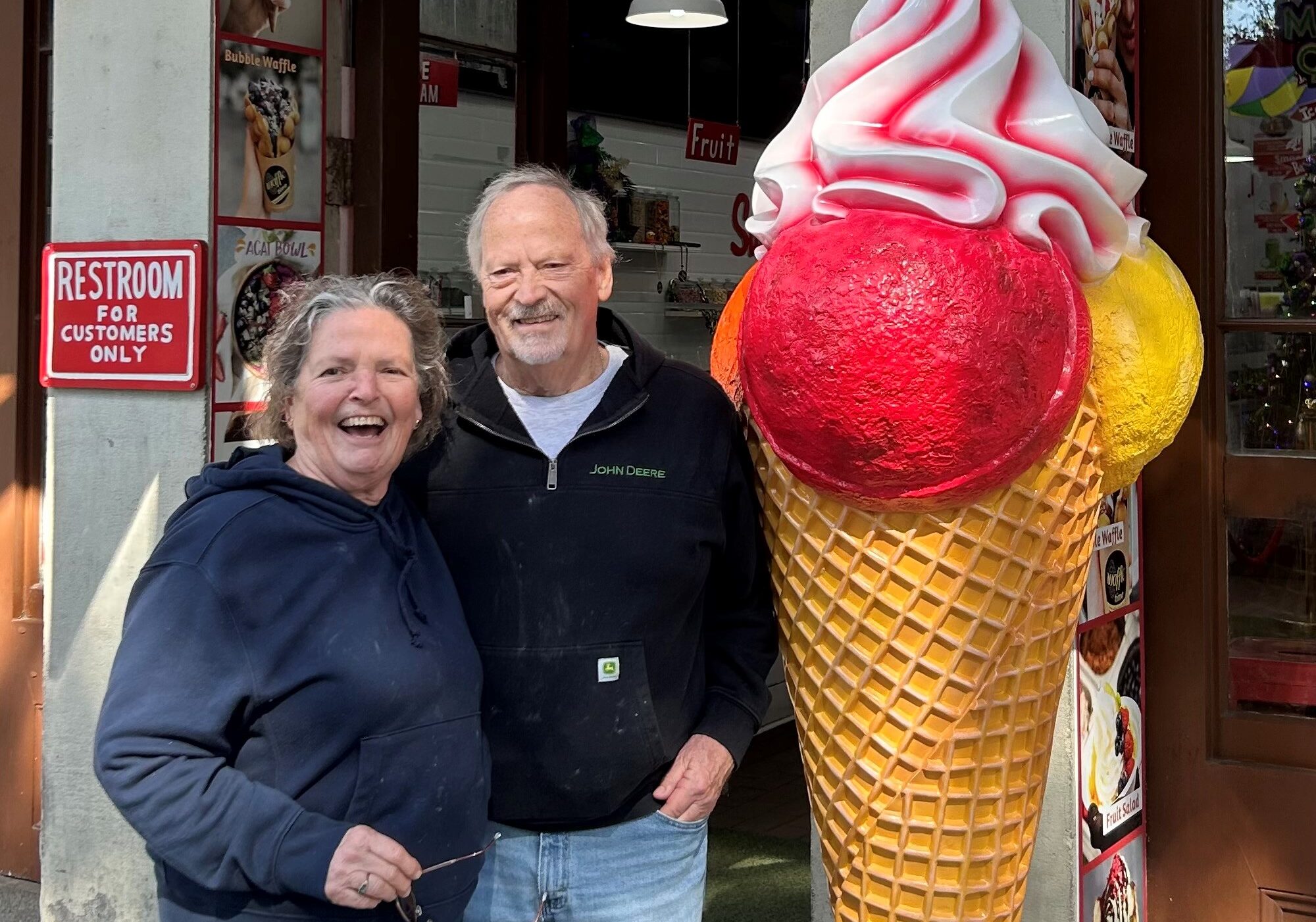 Two people stand outside a storefront next to a large decorative ice cream cone statue with pink and red scoops, holding glasses and wearing casual clothing.