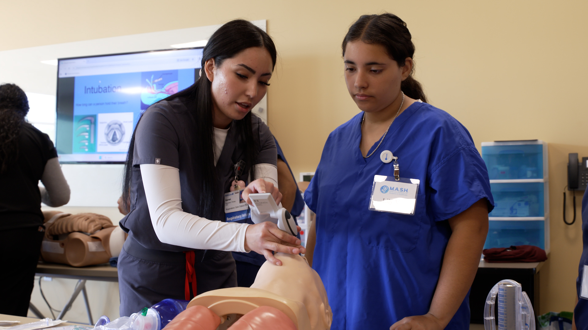 Instructor demonstrates proper use of a handheld airway device on a medical training mannequin while another participant in scrubs observes closely in a classroom setting, with a screen in the background showing intubation visuals.